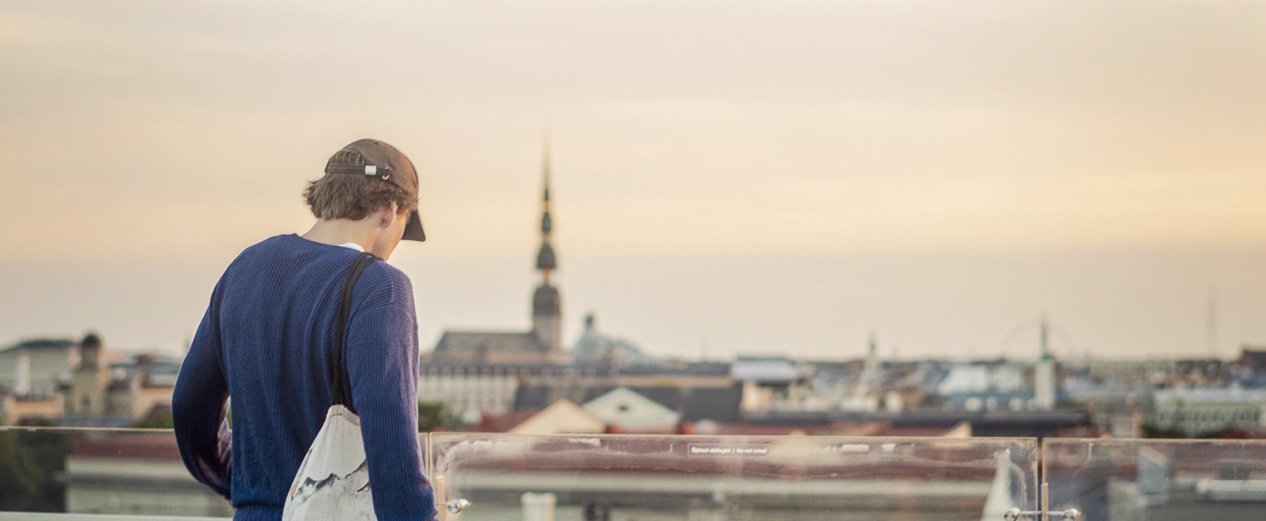 Jauns vīrietis stāv uz CENTRUS ēkas jumta, skatoties uz Rīgas panorāmu ar torņiem un jumtiem pilsētas centrā.